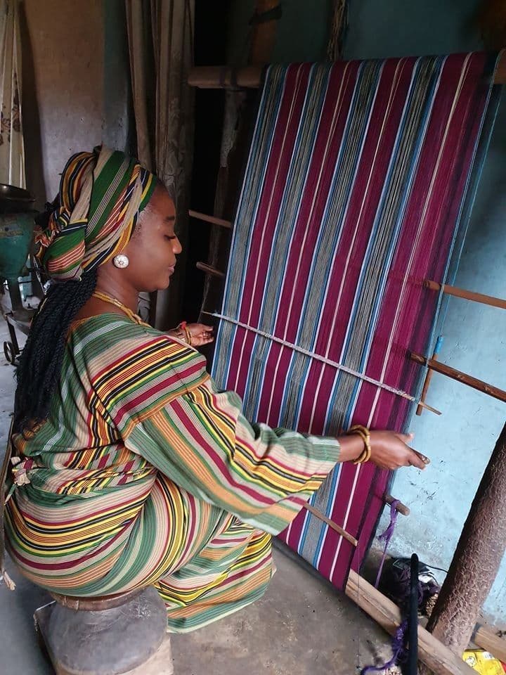 Woman in striped clothing and headwrap weaves colorful fabric on a traditional vertical loom.
